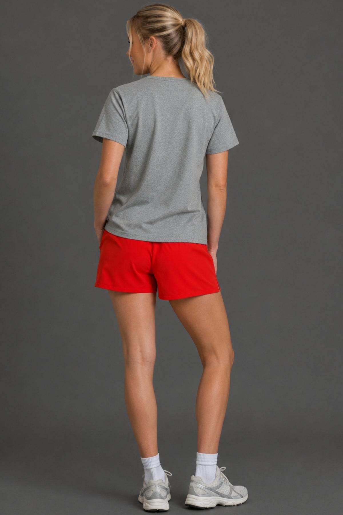 A woman shown from the back wearing a heather gray t-shirt, bright red athletic shorts, white socks, and gray running shoes against a solid dark gray studio background. || Light Heather Gray