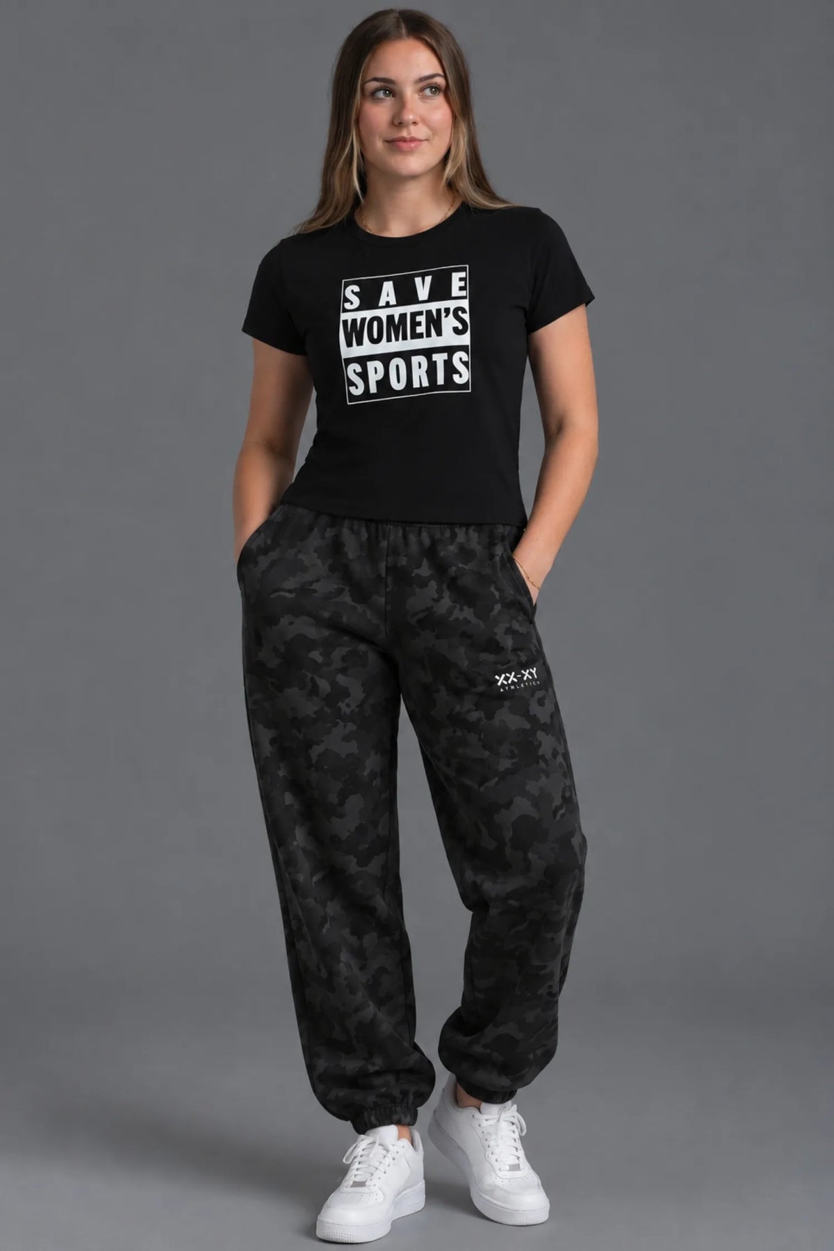 Female model wearing a cropped black Save Women’s Sports graphic tee with black camo sweatpants and white sneakers.