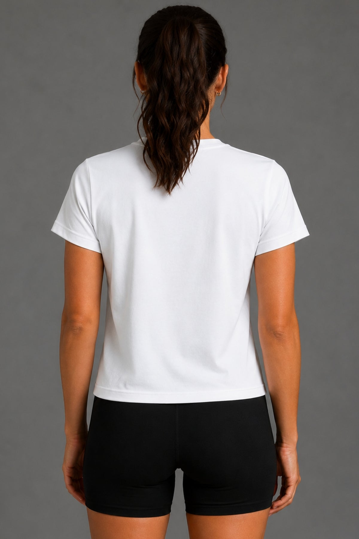 A woman viewed from the back wearing a plain white short-sleeve T-shirt and black fitted shorts against a solid gray studio background.
