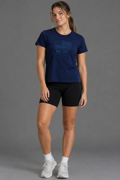 Female model wearing a navy tonal American flag graphic tee with black bike shorts, white socks, and gray sneakers. || Navy