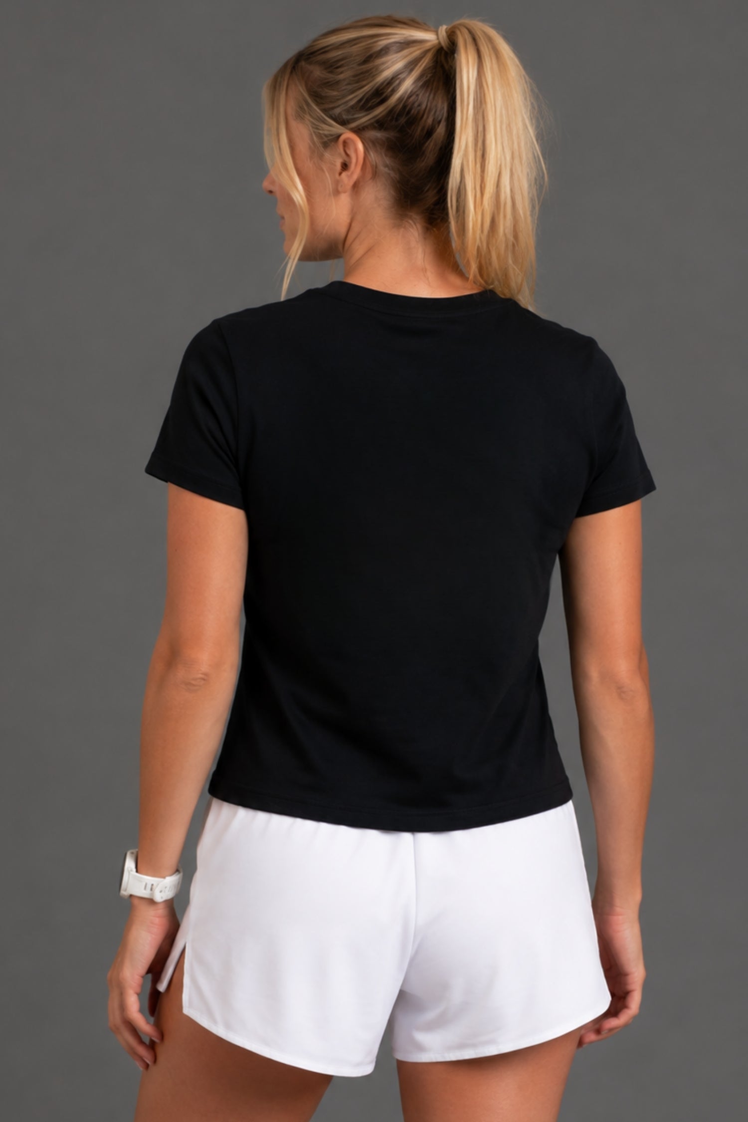 A woman shown from the back wearing a black short-sleeve crewneck t-shirt and white athletic shorts, standing against a solid dark gray studio background. || Black