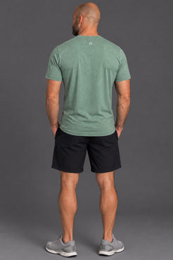 A man viewed from behind wearing a heathered sage green athletic t-shirt, black athletic shorts, and gray sneakers, standing against a solid gray studio background. || Sage Heather