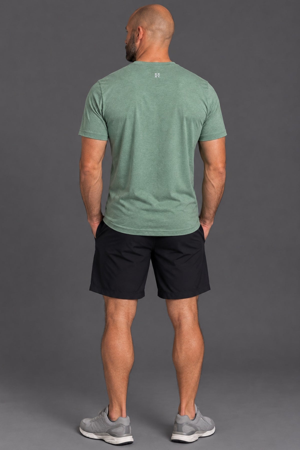 A man viewed from behind wearing a heathered sage green athletic t-shirt, black athletic shorts, and gray sneakers, standing against a solid gray studio background. || Sage Heather