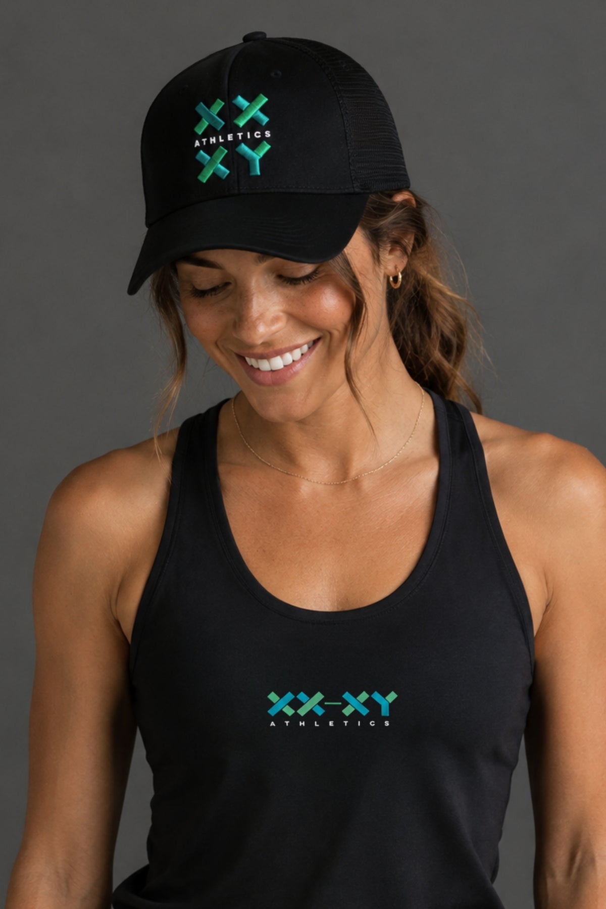 A woman wearing a black trucker hat and matching black tank top, both featuring a green and blue 'XXX-XY Athletics' logo on the front, against a solid dark gray background. || Black with Color Logo