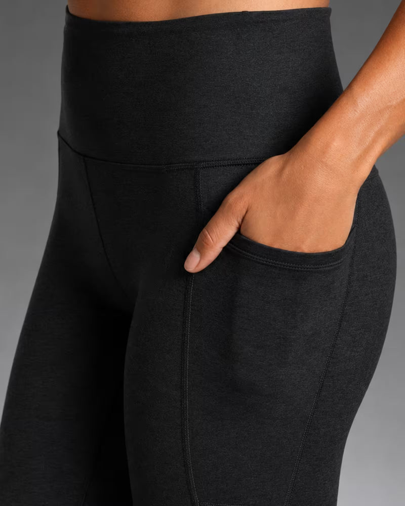 Close-up of a person wearing black high-waisted leggings with a side pocket, showing the waistband, hip seam, and hand in the pocket against a gray studio background.