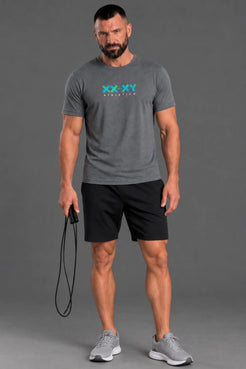 A muscular man wearing a heather gray t-shirt with a blue and green 'XX-XY ATHLETICS' logo on the chest, paired with black athletic shorts and gray sneakers, holding a jump rope against a solid gray studio background. || Dark Heather Gray