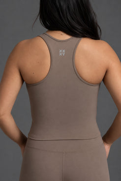 Rear view of a woman wearing a taupe brown athletic tank top and matching leggings against a dark gray studio background. || Mocha