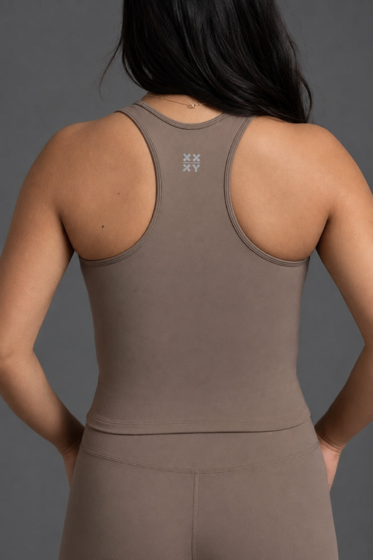 Rear view of a woman wearing a taupe brown athletic tank top and matching leggings against a dark gray studio background. || Mocha