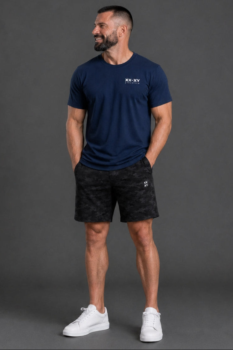 Man wearing a navy blue t-shirt and black shorts with a logo, standing against a gray background