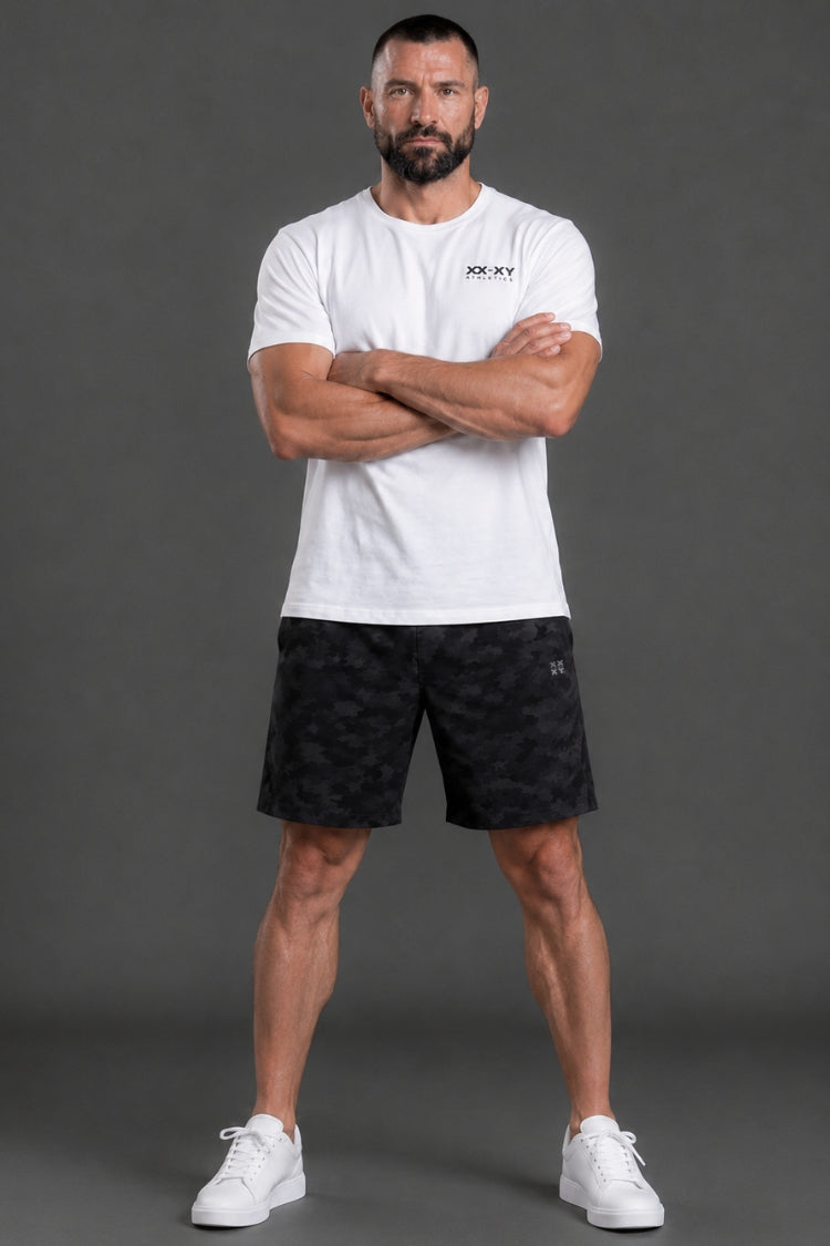 Man wearing a white t-shirt and black shorts with a brand logo, standing against a gray background || White