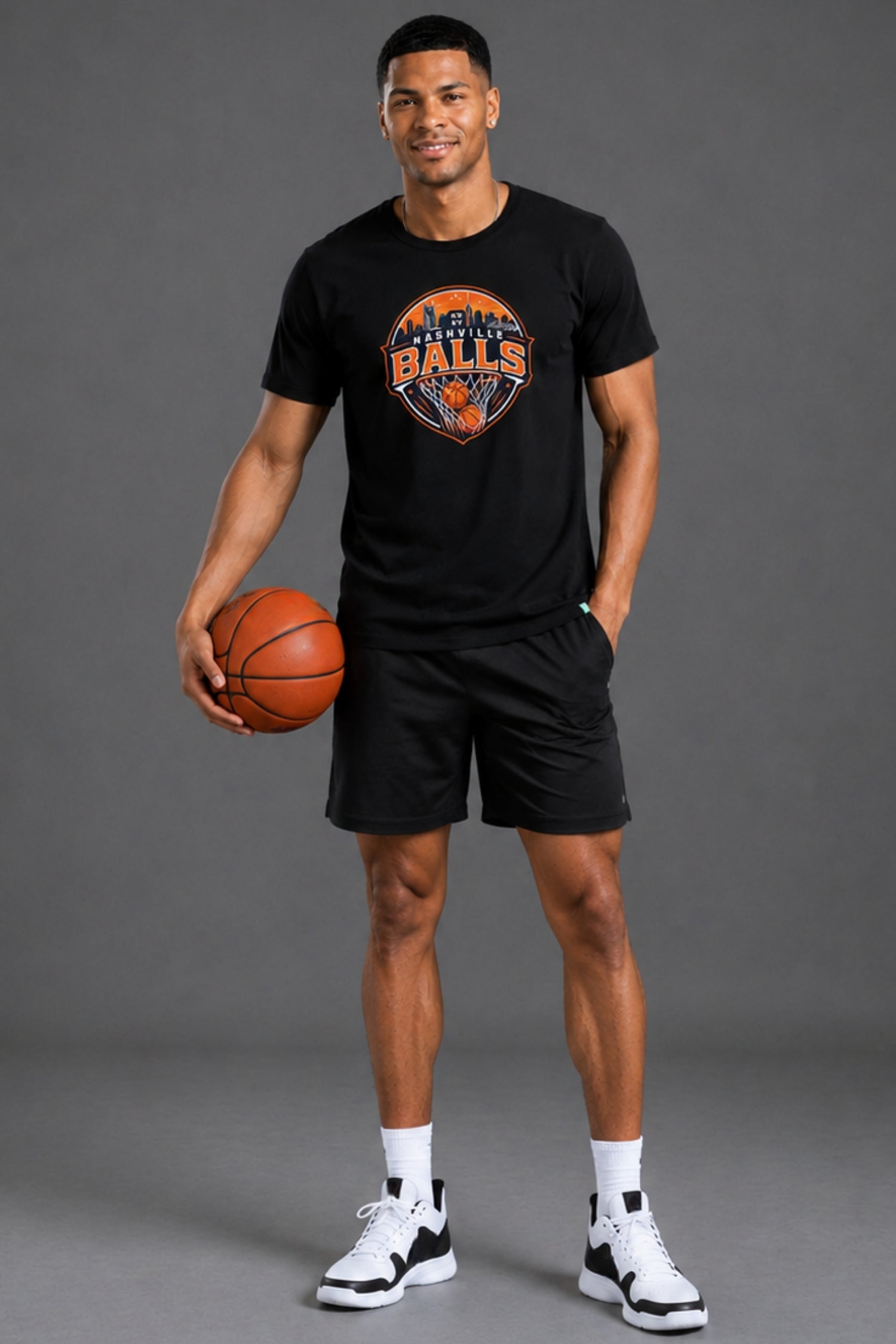 A man modeling a black short-sleeve graphic t-shirt with an orange and blue Phoenix Suns logo, paired with black athletic shorts, white socks, and black-and-white sneakers, holding a basketball against a solid gray studio background.