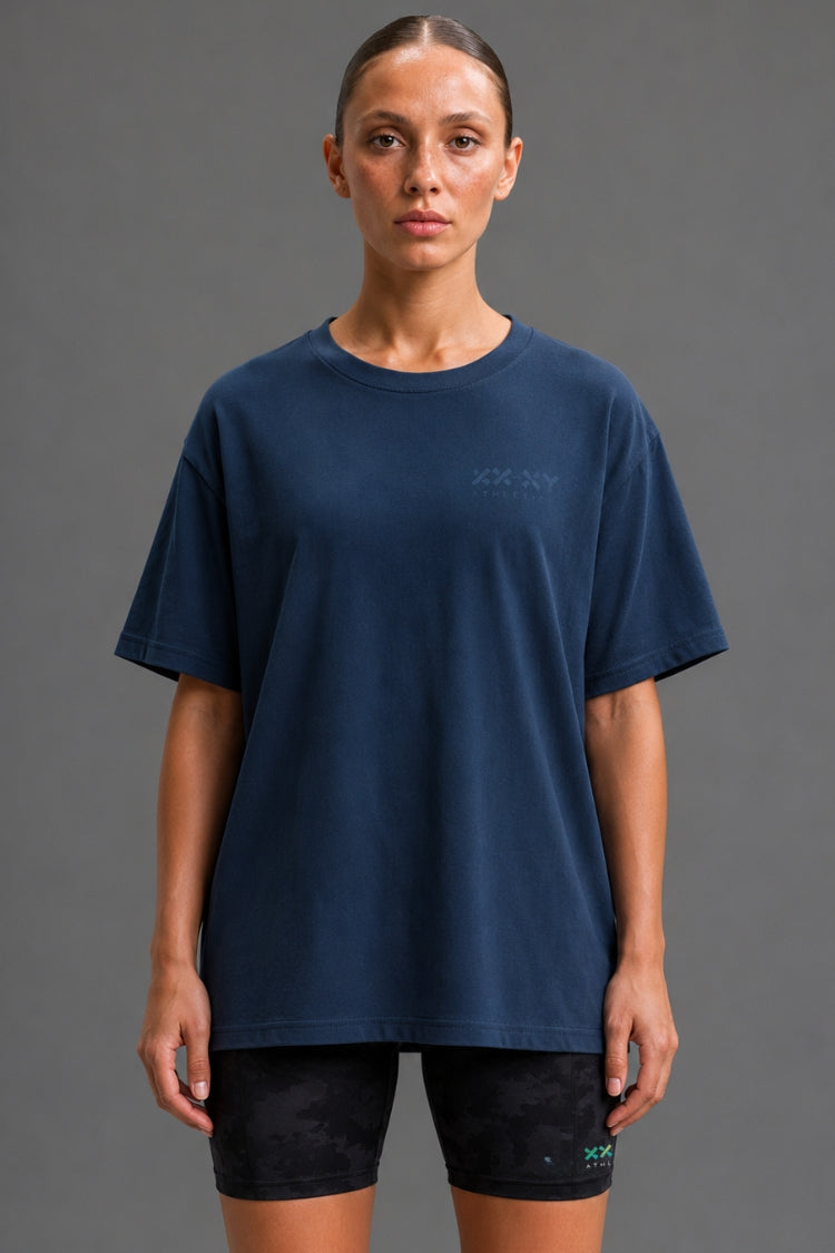 A woman modeling an oversized navy blue t-shirt with a subtle tonal logo on the left chest, paired with black athletic shorts, against a solid gray studio background.