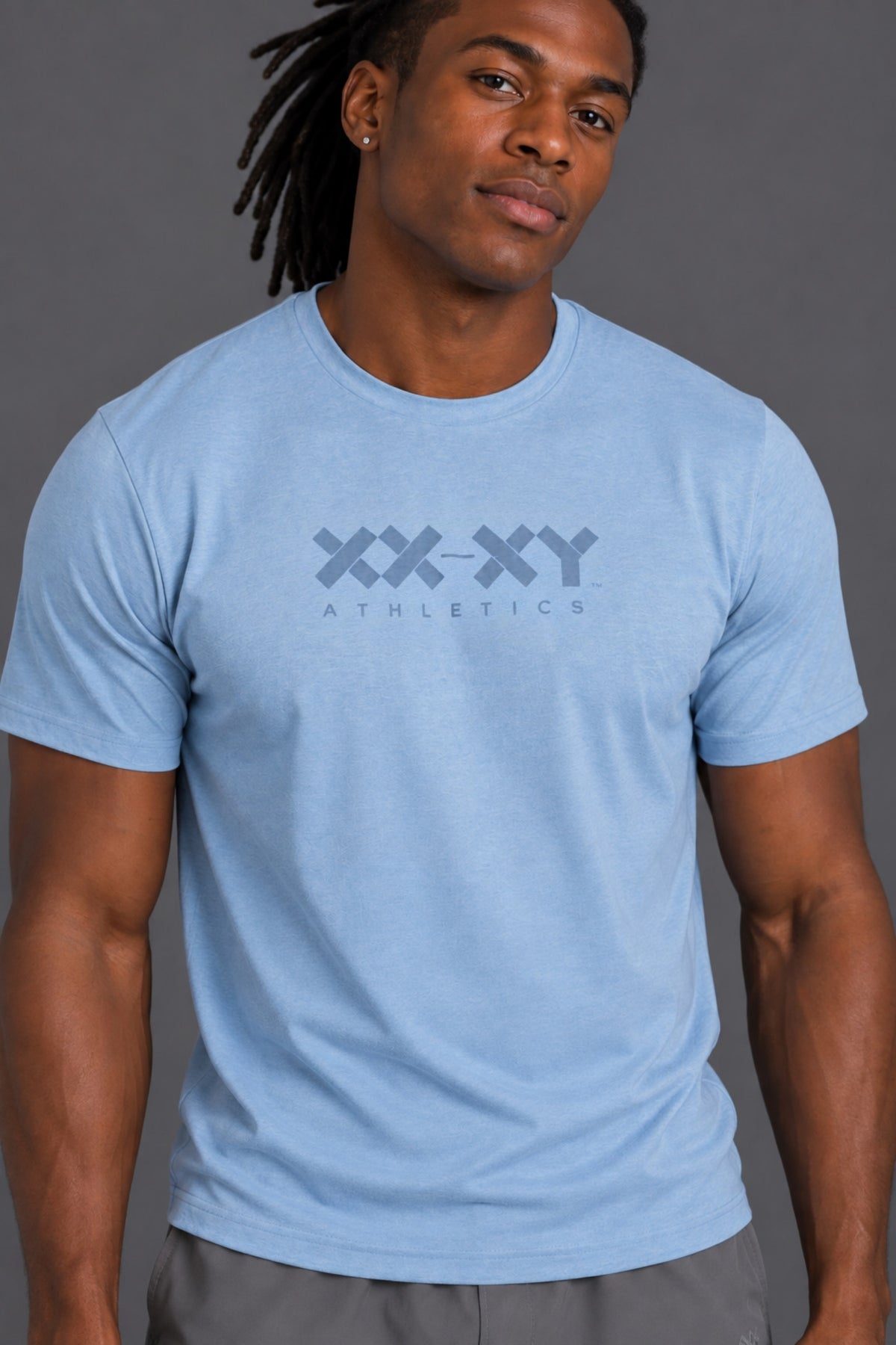 A man wearing a light blue crewneck t-shirt with a dark gray 'XX-XY ATHLETICS' logo printed on the chest, set against a solid gray studio background.