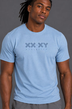 A man wearing a light blue crewneck t-shirt with a dark gray 'XX-XY ATHLETICS' logo printed on the chest, set against a solid gray studio background. || Cerulean Blue Heather
