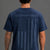 A man viewed from the back wearing a navy blue t-shirt with a distressed American flag graphic and the words 'SAVE WOMEN'S SPORTS' across the upper back, paired with red athletic shorts, white socks, and gray sneakers, against a solid gray studio background. || Navy