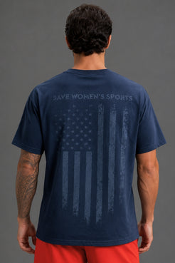 A man viewed from the back wearing a navy blue t-shirt with a distressed American flag graphic and the words 'SAVE WOMEN'S SPORTS' across the upper back, paired with red athletic shorts, white socks, and gray sneakers, against a solid gray studio background. || Navy