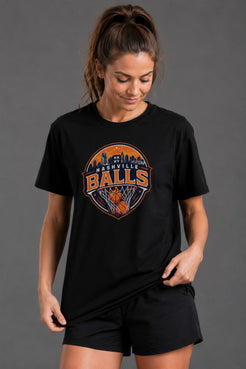 A woman wearing a black short-sleeve graphic T-shirt and black shorts against a solid gray studio background. The shirt features a circular 'Tennessee Balls' basketball graphic in orange, white, and blue on the chest. || Black