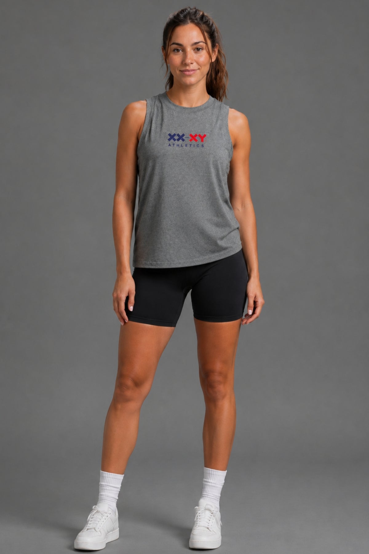 A woman modeling a heather gray sleeveless athletic tank top with a red and blue 'XX XV ATHLETICS' logo on the chest, paired with black athletic shorts, white socks, and white sneakers, against a solid gray studio background.