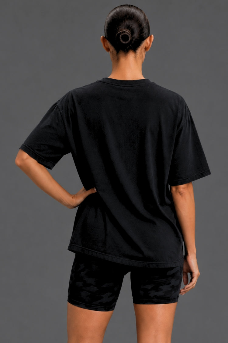A woman shown from the back wearing a black oversized short-sleeve t-shirt and black patterned athletic shorts against a dark gray studio background.