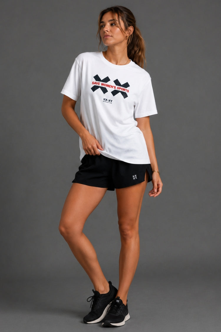 A woman modeling a white short-sleeve athletic T-shirt with a black and red XX Sports logo on the chest, paired with black shorts and black sneakers, against a solid gray studio background.
