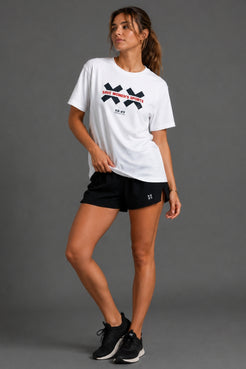 A woman modeling a white short-sleeve athletic T-shirt with a black and red XX Sports logo on the chest, paired with black shorts and black sneakers, against a solid gray studio background. || White