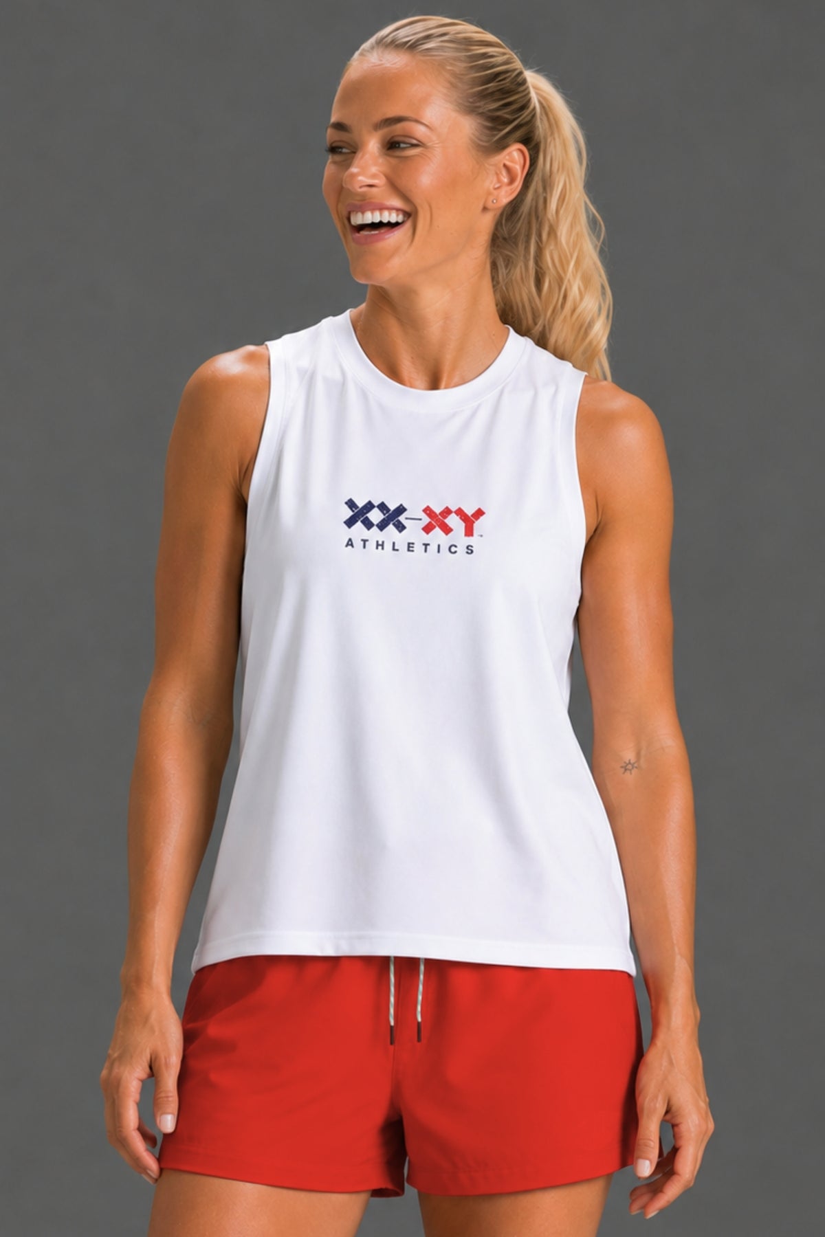 A woman wearing a white sleeveless athletic tank top with a red and navy 'XX-XY Athletics' logo on the chest, paired with red athletic shorts, against a solid gray studio background. || White