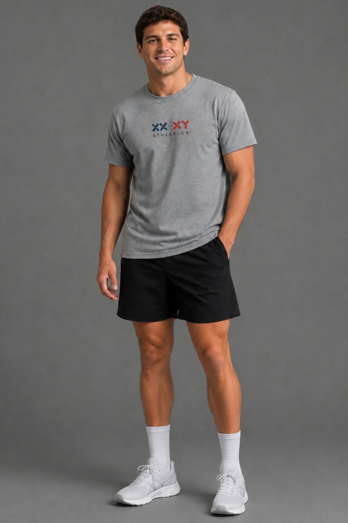 A man modeling a heather gray short-sleeve t-shirt with a 'XX-XY Athletics' logo on the chest, paired with black athletic shorts, white socks, and white sneakers, against a solid gray studio background. || Heather Gray