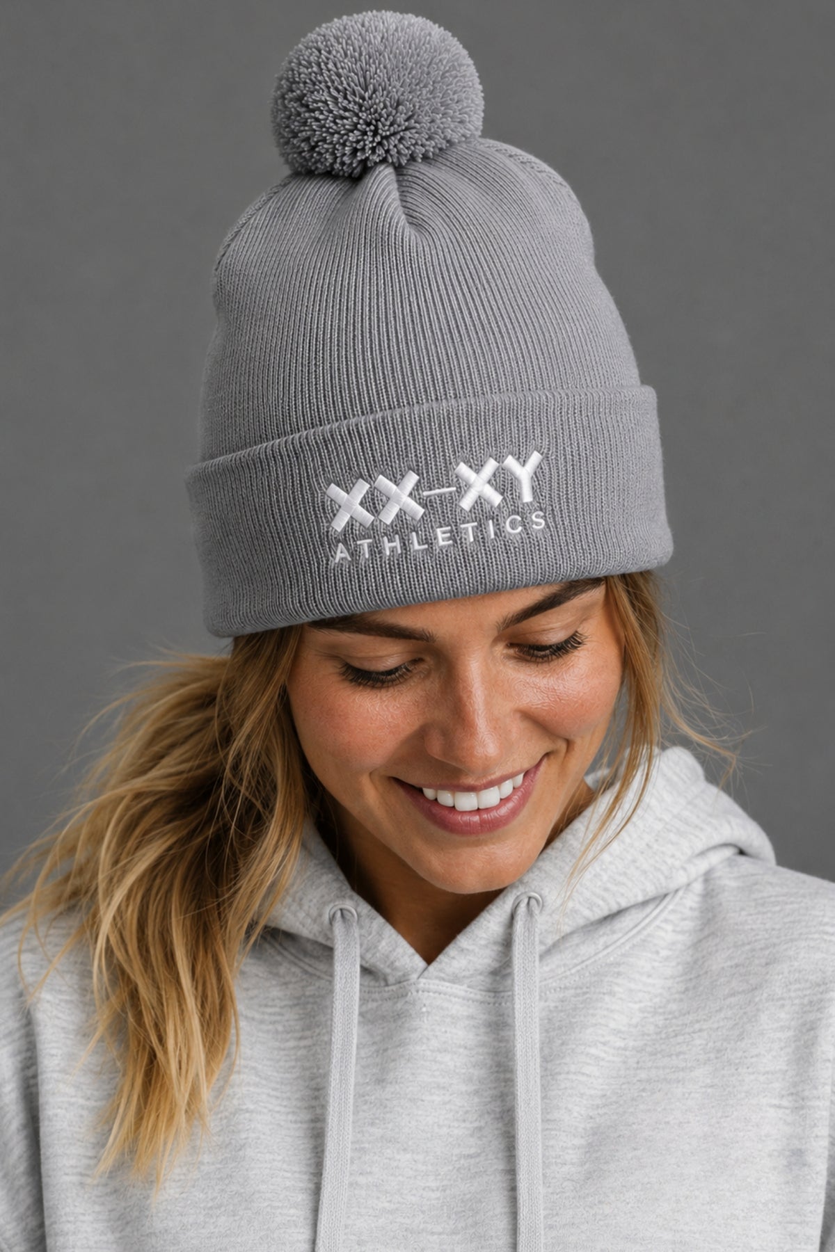 A woman wearing a gray ribbed knit pom-pom beanie with 'XX-XY ATHLETICS' embroidered in white on the front cuff, paired with a light gray hoodie against a dark gray background.