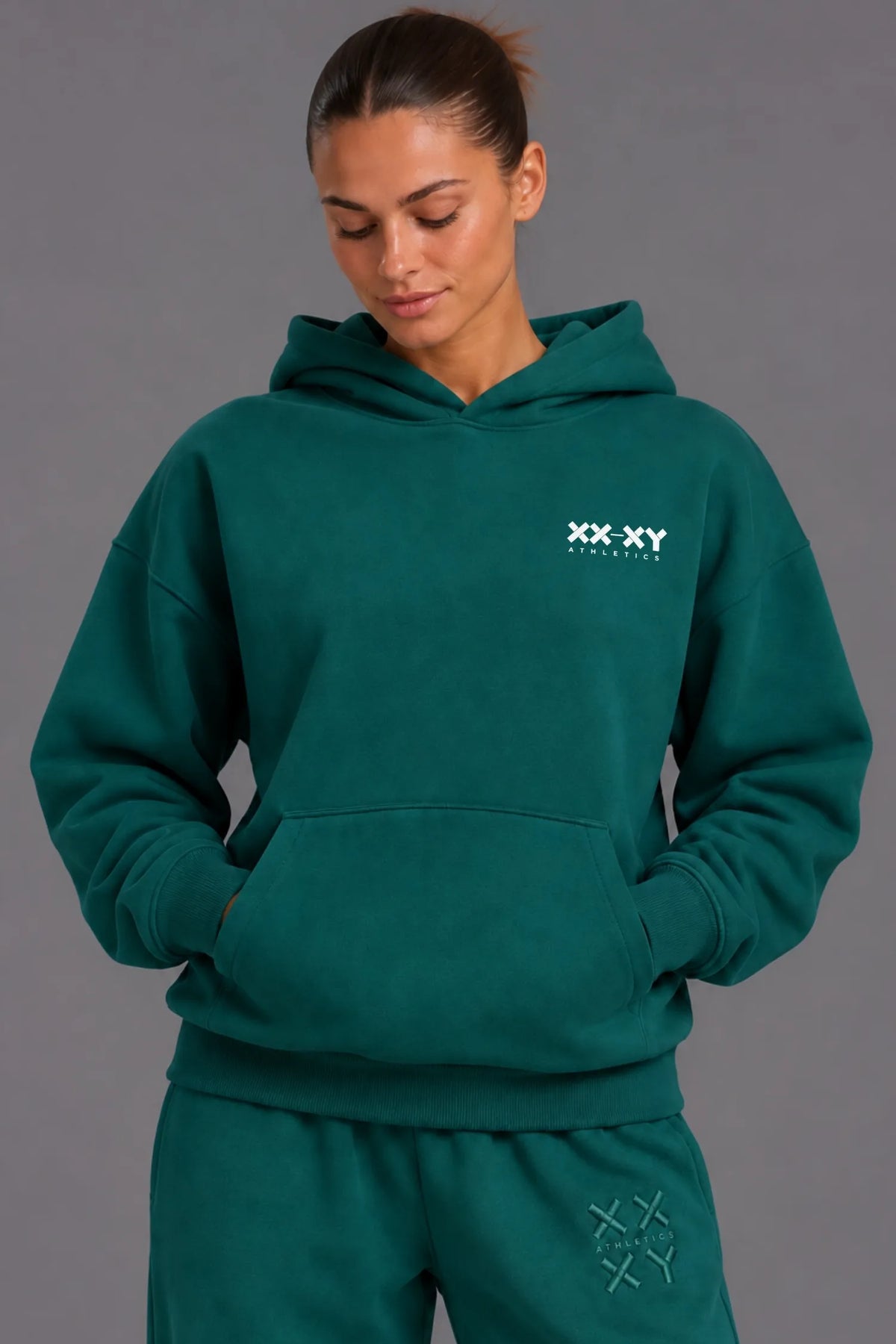 Woman wearing a teal XX-XY Athletics hoodie and matching sweatpants against a gray studio background. || Pine