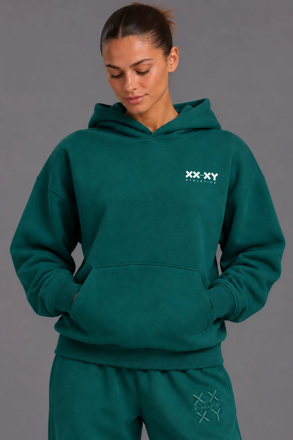 Woman wearing a teal XX-XY Athletics hoodie and matching sweatpants against a gray studio background. || Pine