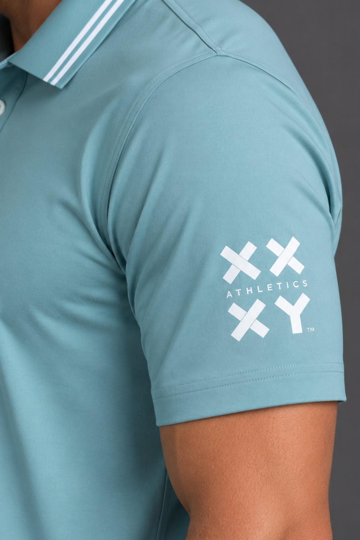 Close-up of a person wearing a light blue polo shirt with a white XX Athletics logo on the sleeve and a white stripe detail on the collar, set against a dark gray background. || Underseas