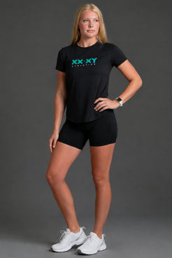 A woman modeling a black short-sleeve athletic T-shirt with a teal and white 'XX XY' logo on the chest, paired with black fitted shorts and white sneakers, against a dark gray studio background. || Black