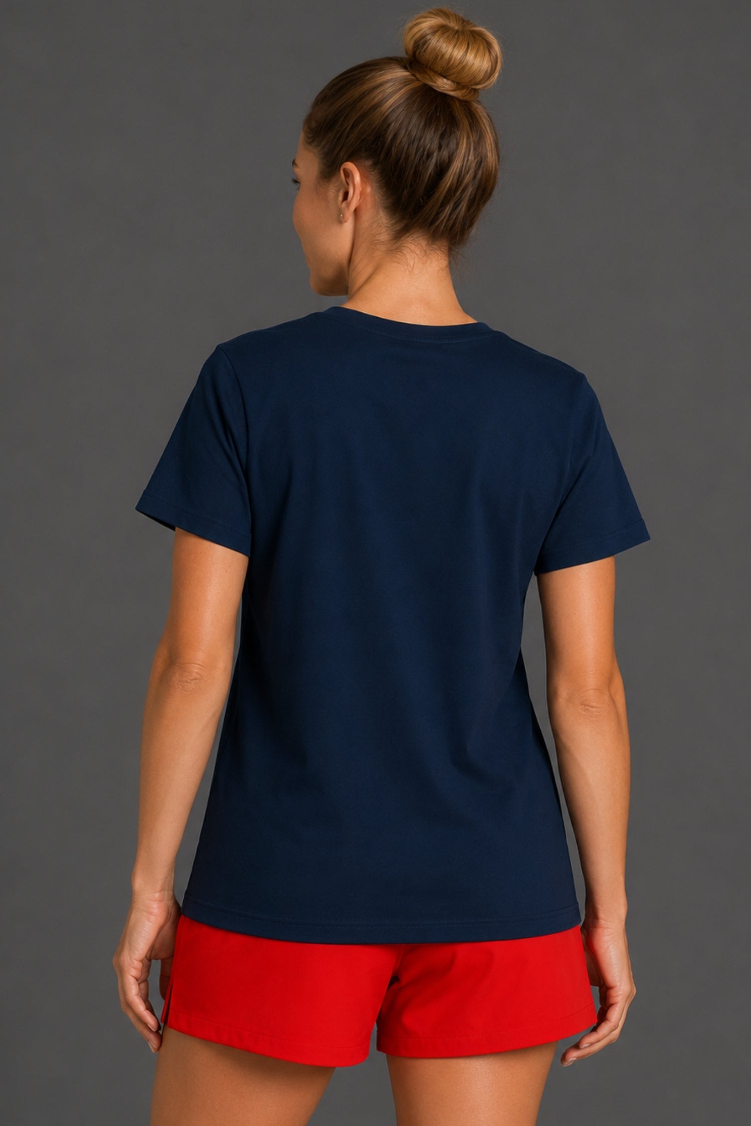 A woman shown from the back wearing a navy blue short-sleeve t-shirt and bright red shorts against a dark gray studio background.
