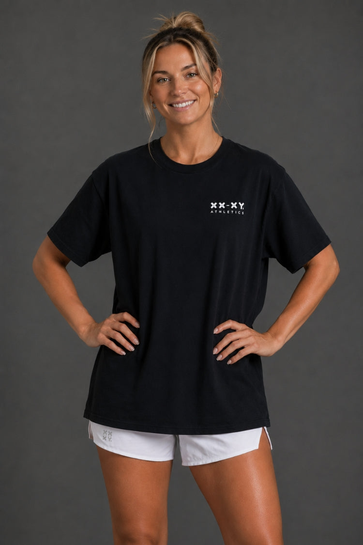 A woman modeling a black short-sleeve t-shirt with a small white XX-XY Athletics logo on the left chest, paired with white athletic shorts, against a dark gray studio background.