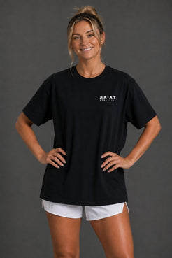 A woman modeling a black short-sleeve t-shirt with a small white XX-XY Athletics logo on the left chest, paired with white athletic shorts, against a dark gray studio background. || Black
