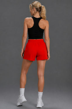 A woman shown from the back wearing bright red athletic shorts, a black sports bra, white socks, and white sneakers against a solid gray studio background.|| Red