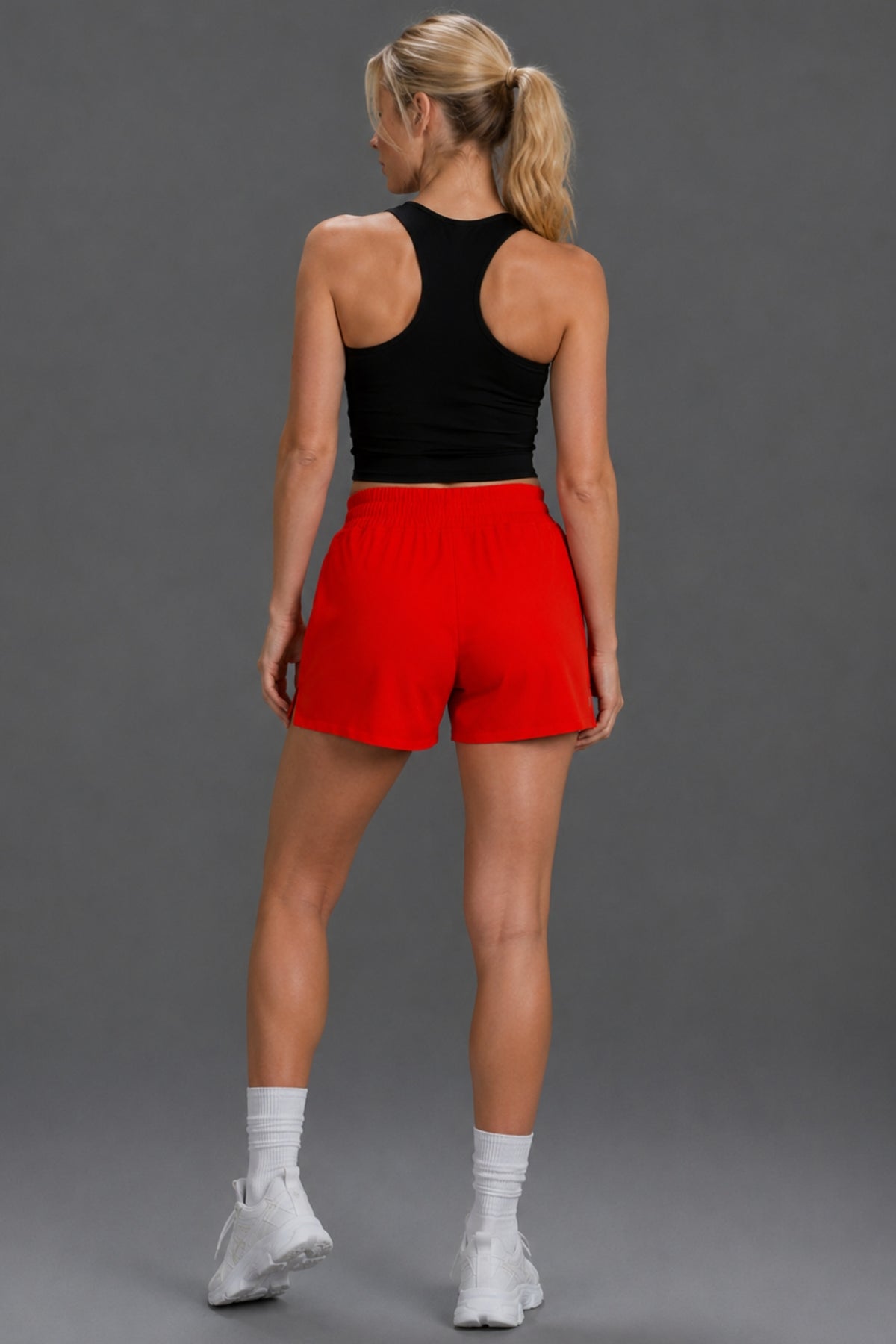 A woman shown from the back wearing bright red athletic shorts, a black sports bra, white socks, and white sneakers against a solid gray studio background.|| Red