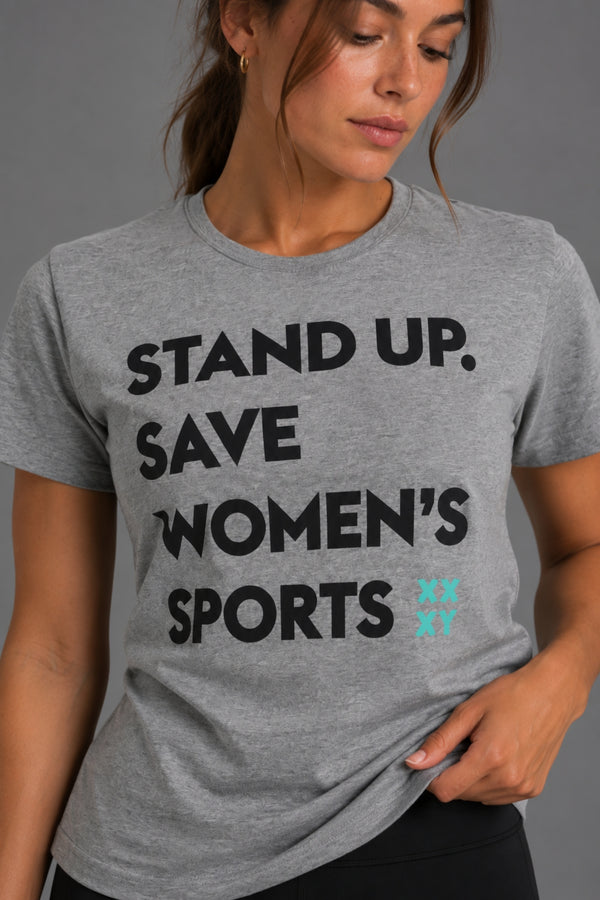 A woman wearing a heather gray graphic T-shirt with the text 'STAND UP. SAVE WOMEN'S SPORTS' in black and a small turquoise logo, photographed against a plain gray studio background. || Heather Gray