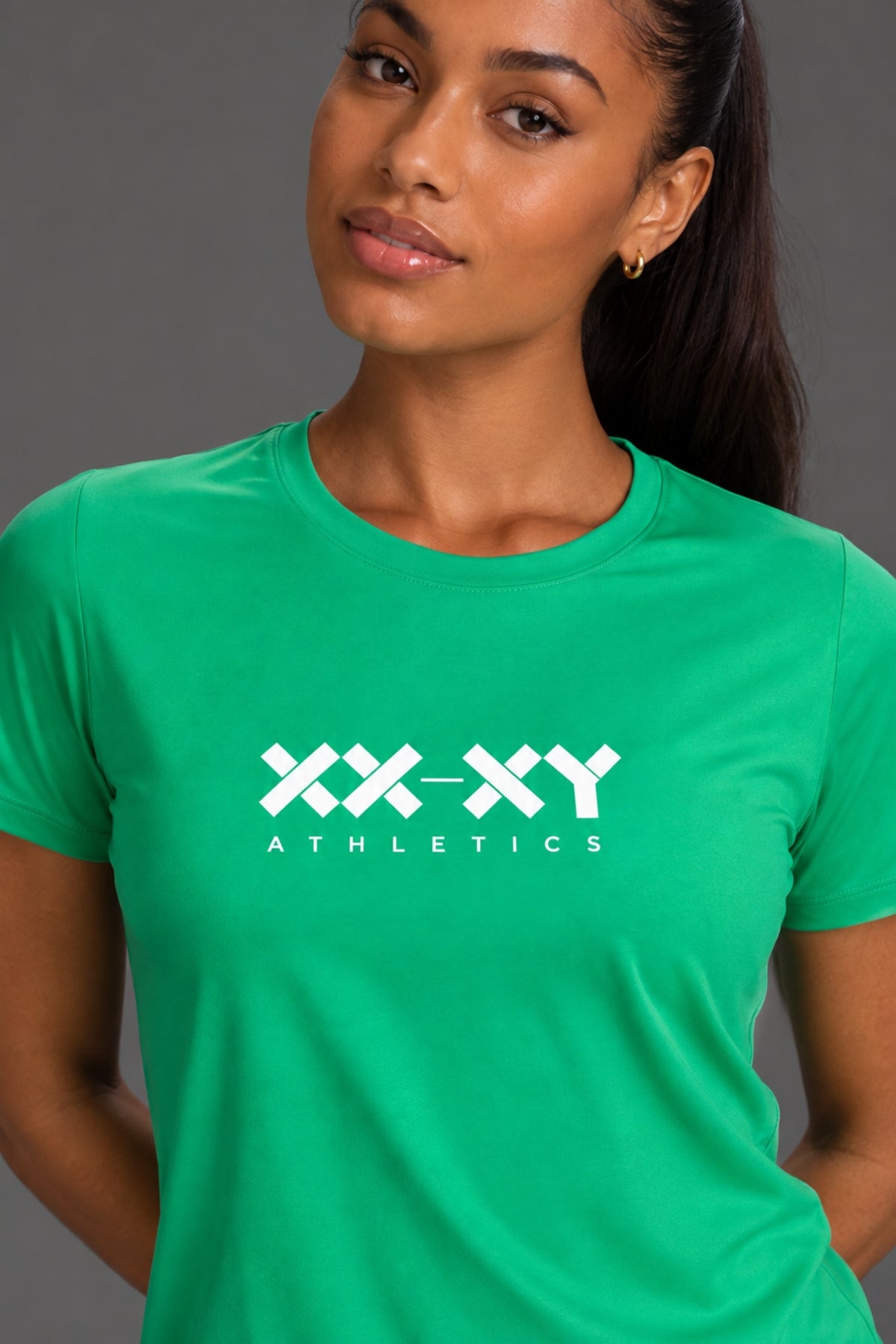 A woman wearing a bright green short-sleeve crewneck t-shirt with a white 'XX-XY ATHLETICS' logo printed on the chest, set against a solid gray studio background.