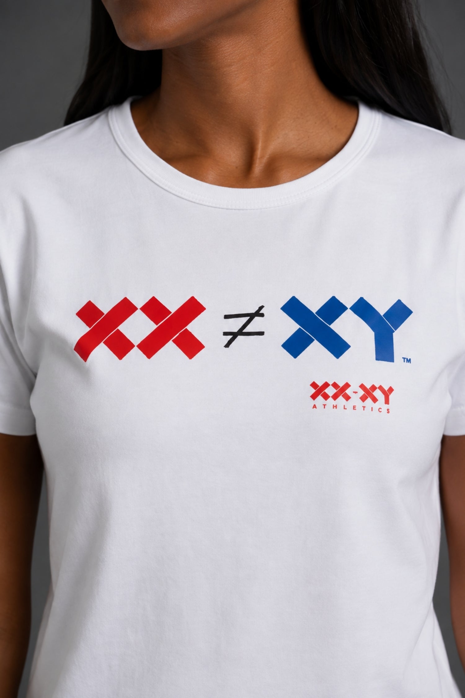 A close-up of a white crewneck t-shirt worn by a model. The shirt features a graphic print on the chest reading 'XX ≠ XY' in red and blue, with 'XX-XY Athletics' printed below in red. || White