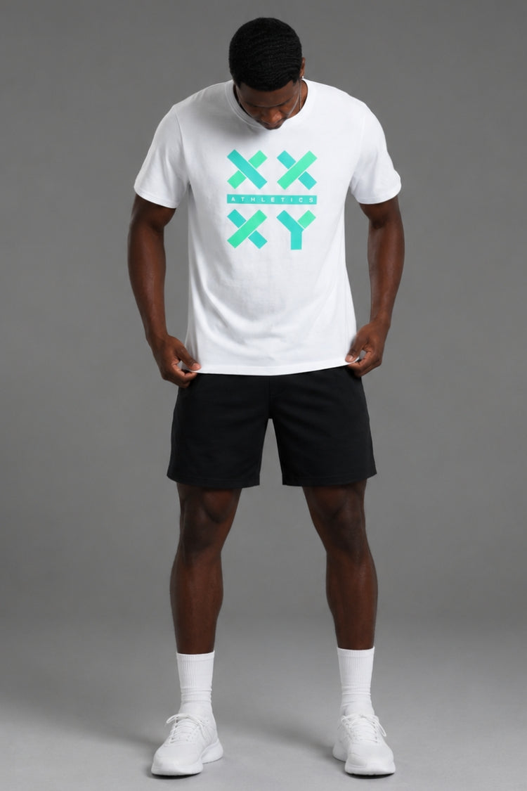 A man wearing a white crewneck t-shirt with a mint green XXYY logo on the chest, paired with black athletic shorts, white socks, and white sneakers, standing against a solid gray studio background.