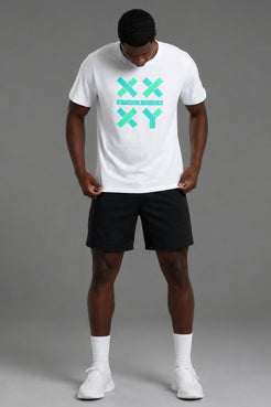 A man wearing a white crewneck t-shirt with a mint green XXYY logo on the chest, paired with black athletic shorts, white socks, and white sneakers, standing against a solid gray studio background. || White