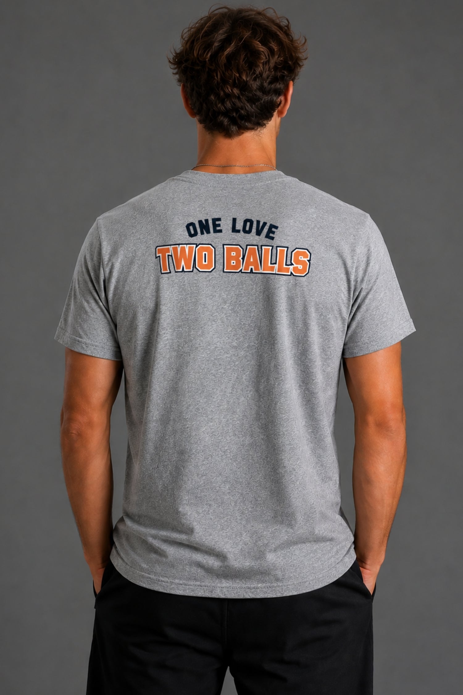 A man viewed from behind wearing a heather gray t-shirt with 'ONE LOVE TWO BALLS' printed on the back in navy and orange, paired with black pants against a solid gray studio background. || Heather Gray