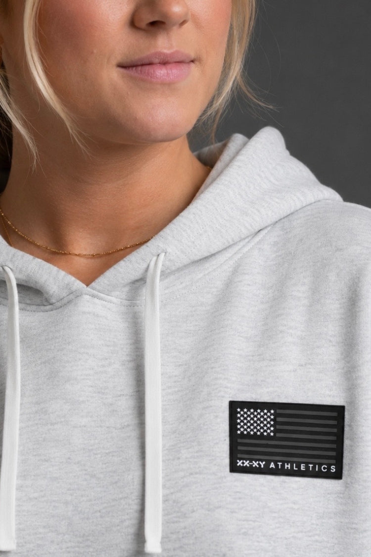 Person wearing a gray hoodie with a logo on the chest against a dark background || Light Heather Gray
