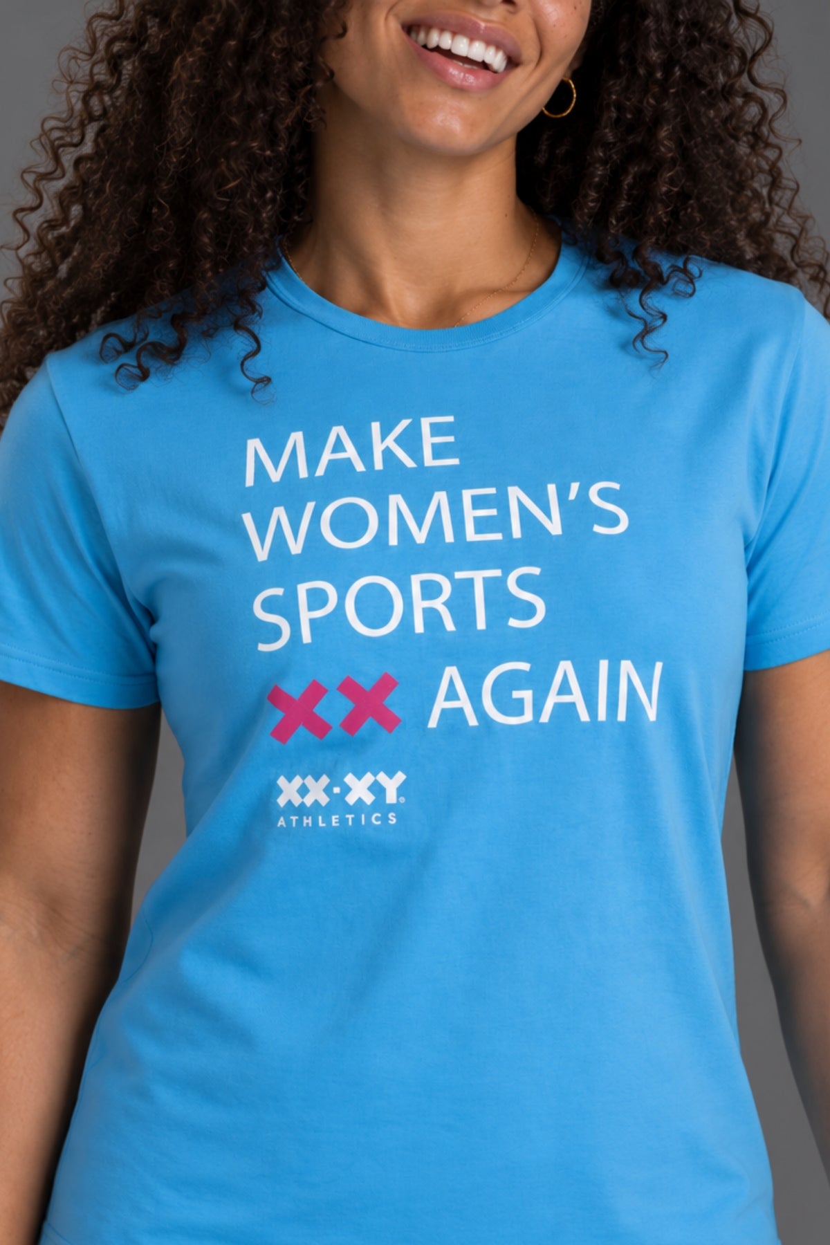 A woman wearing a bright blue graphic t-shirt with the text 'MAKE WOMEN'S SPORTS XX AGAIN' and the 'XXX-XY Athletics' logo printed on the front.