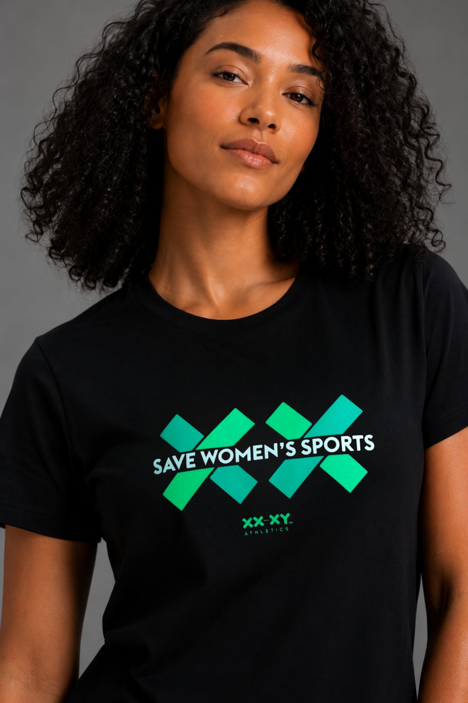 A woman with curly black hair wearing a black short-sleeve t-shirt with a green and white graphic that reads 'SAVE WOMEN'S SPORTS' and 'XX XY'.