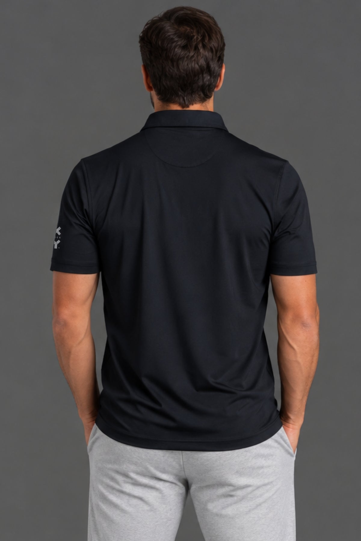A man viewed from behind wearing a black short-sleeve polo shirt and light gray sweatpants, standing against a solid dark gray studio background.