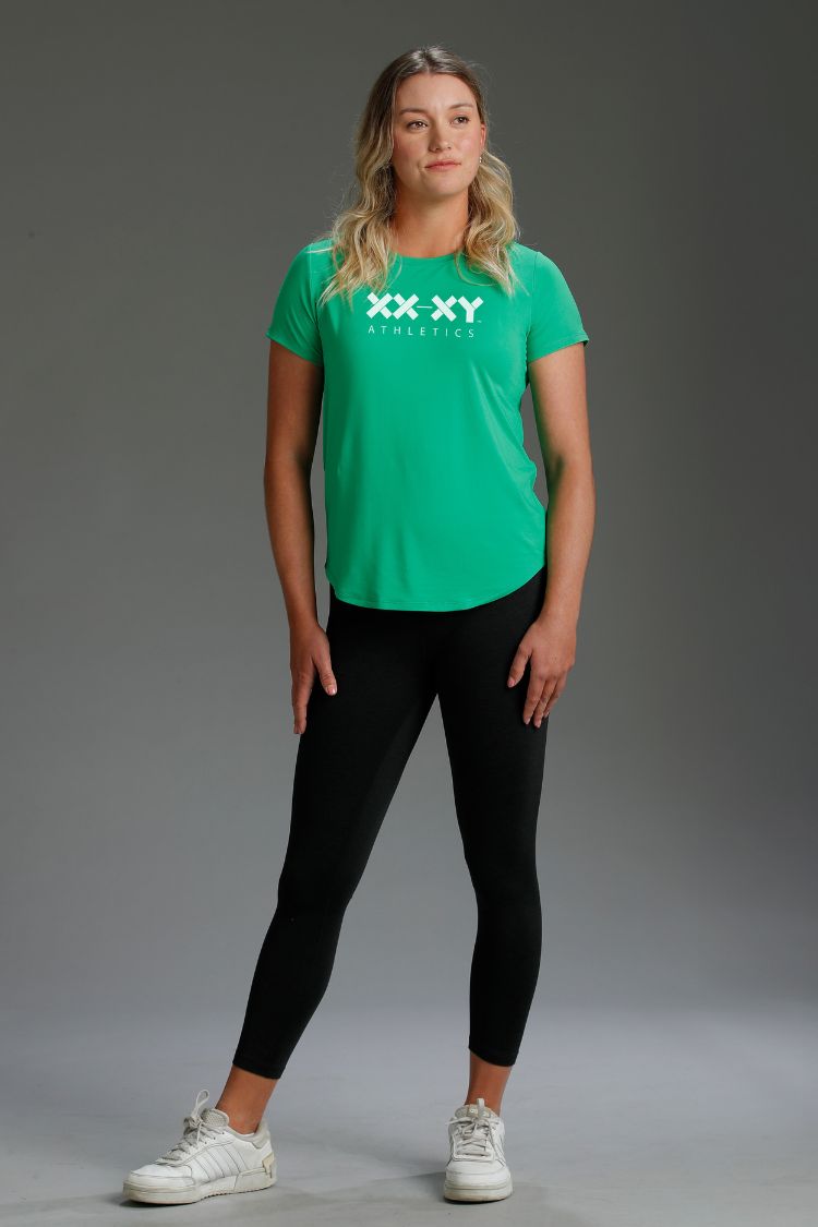 Woman wearing a green t-shirt with a logo and black leggings on a gray background || Kelly Green