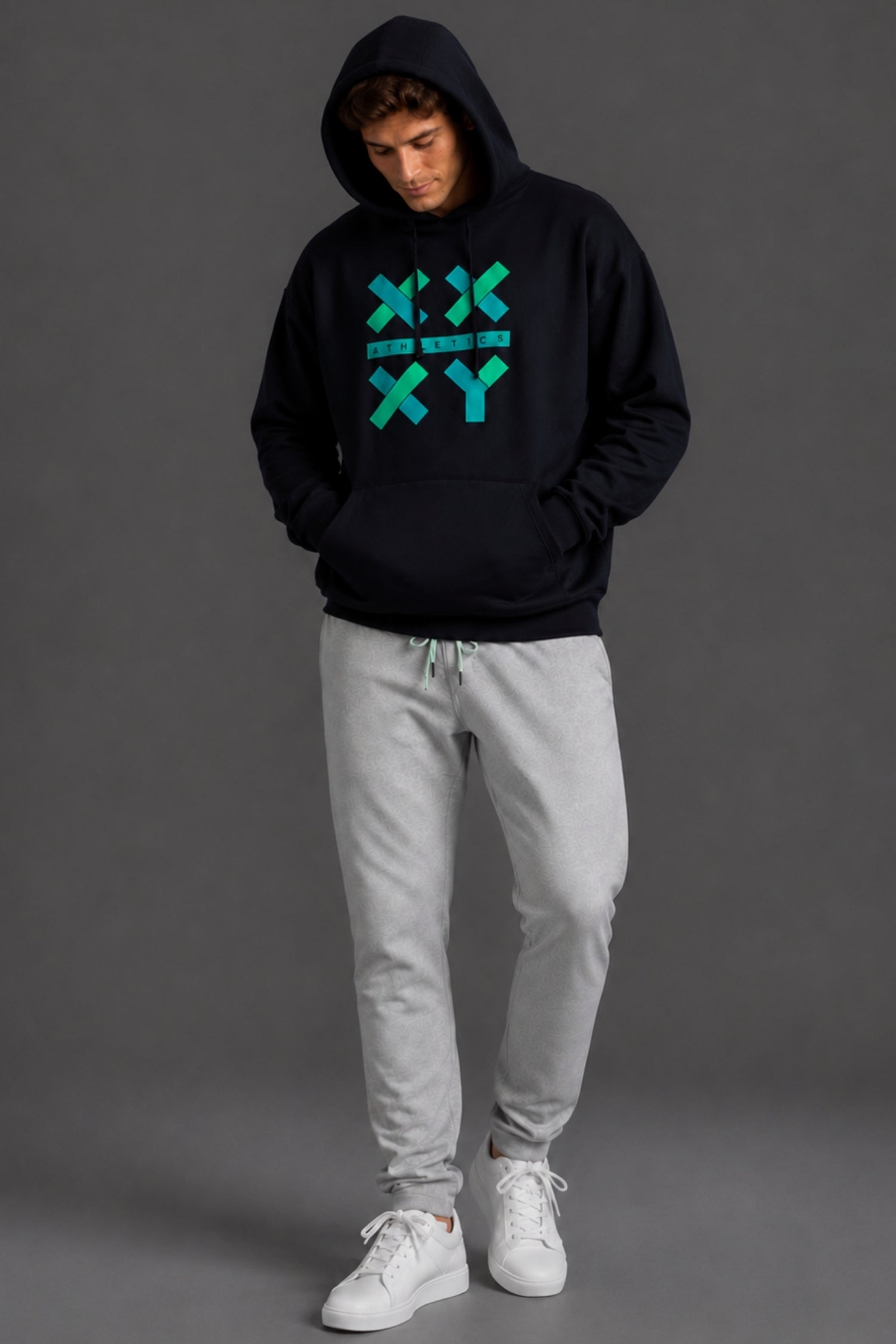A man wearing a black pullover hoodie with a teal geometric 'XY' graphic on the chest, paired with light gray sweatpants and white sneakers, standing against a dark gray studio background.