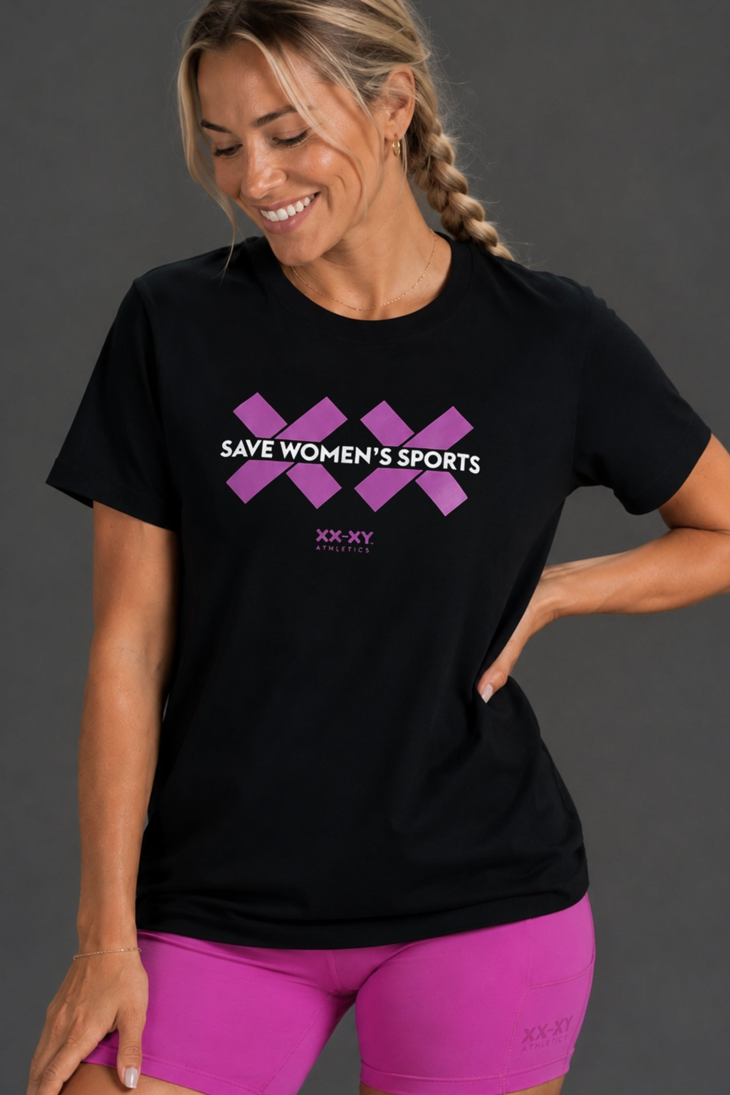 A woman wearing a black short-sleeve t-shirt with a purple graphic that reads 'SAVE WOMEN'S SPORTS' and 'XX-XY SPORTS' underneath, paired with bright pink athletic shorts, against a dark gray studio background.
